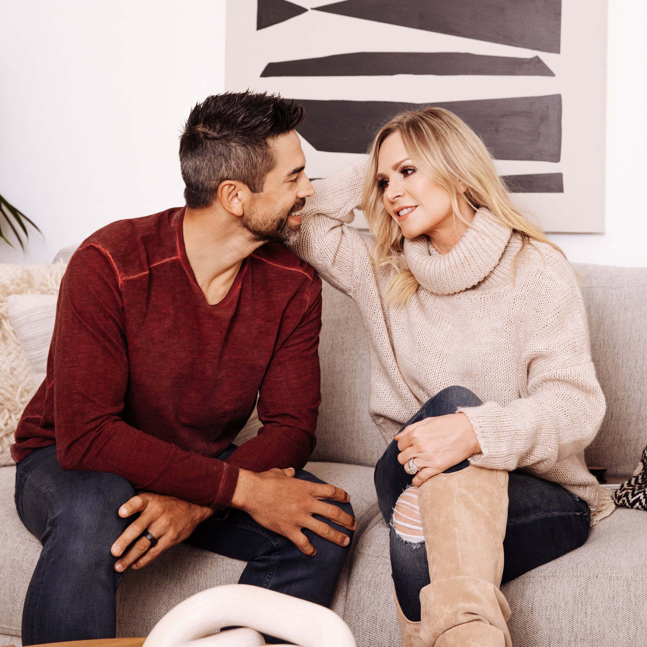Smiling couple sitting on a couch facing each other — woman in beige turtleneck and suede boots, man in burgundy sweater.